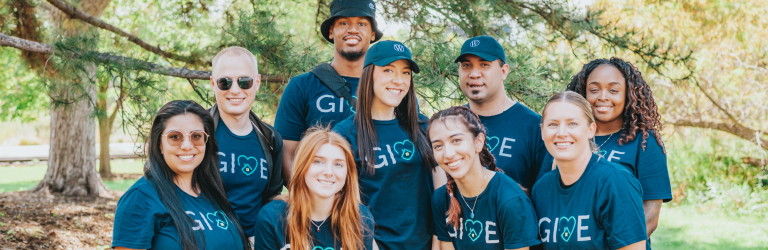 A group of individuals outdoors posing together, wearing matching navy blue shirts with the GIVE logo and a heart symbol. Some individuals wearing navy blue caps with a logo on the front