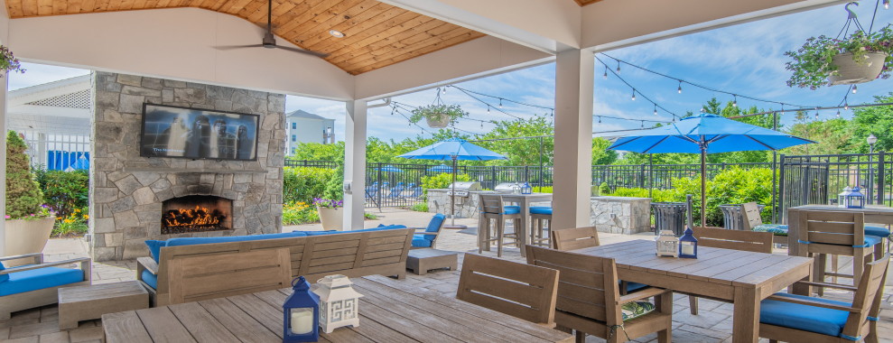 An outdoor patio area with wooden table and chairs in front of a fireplace. The patio area is covered with a roof with wooden paneling held up by white support columns