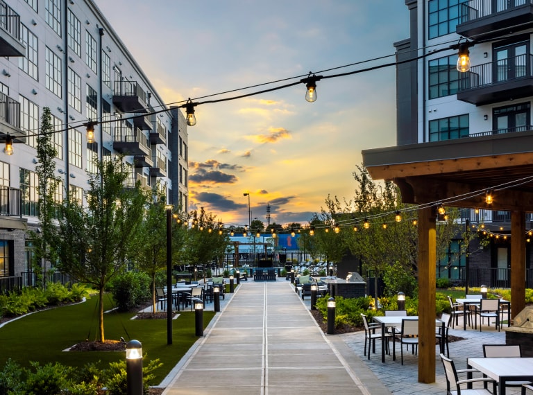 View down a pathway between mid-rise residential buildings at sunset, with outdoor chairs and tables and landscaped gardens