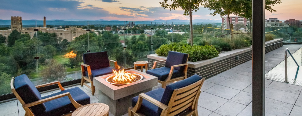 Outdoor seating area on a tiled patio with a fire pit. The background is a green landscape with a city in the distance
