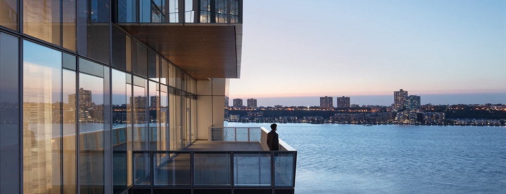 A waterfront cityscape, a person standing on a balcony. The background shows a body of water and several high-rise buildings