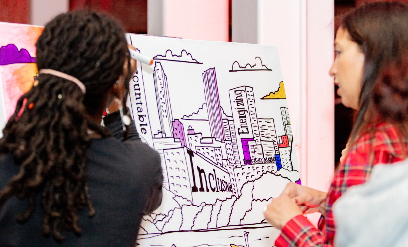 Two individuals working on a large, partially colored illustation. The illustration features a cityscape with buildings and text including the words Energizing and Inclusive