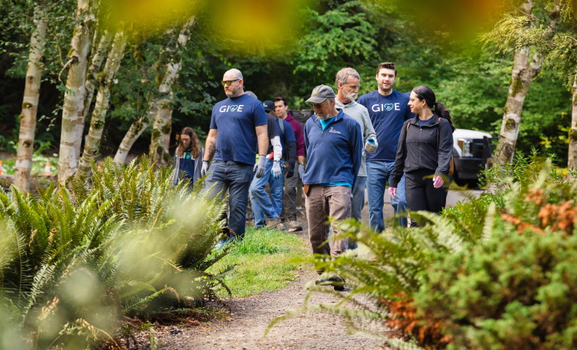 A group of individuals walking through a garden area with trees and shrubs. Some of the individuals are wearing matching navy blue shirts with GIVE logo and heart symbol