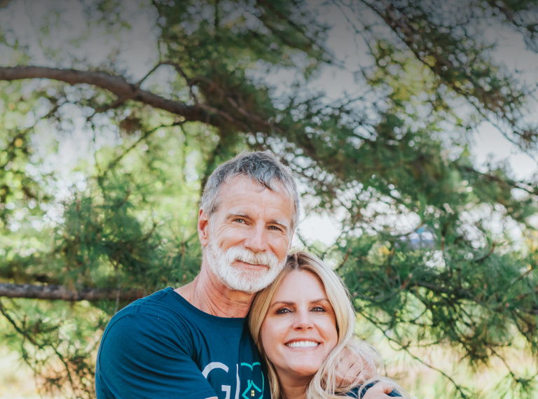 A man with gray hair and beard embracing a woman with long blonde hair, both wearing navy blue shirts with the GIVE logo and heart symbol. They are outdoors with trees and greenery in the background