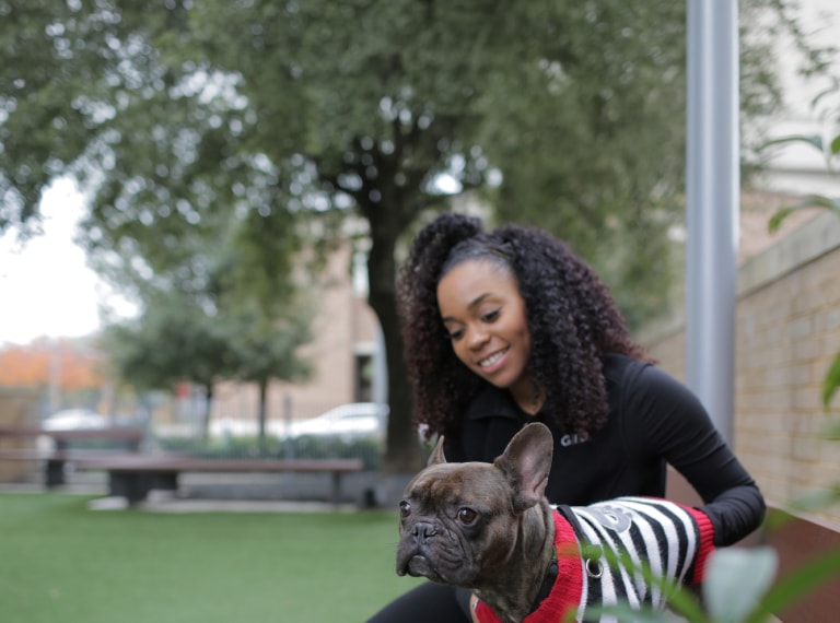 A person sitting on a bench ion the park with a French bulldog wearing a stripy jacket