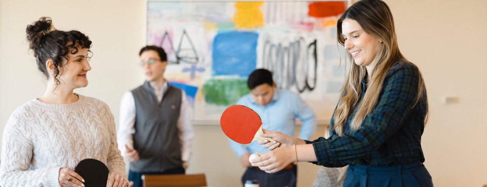 Two individuals holding ping pong paddles and ball. Other people are standing in the blurred background
