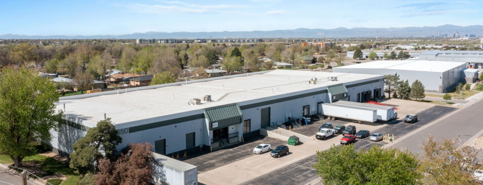 An aerial view of a rectangular building with a flat roof and multiple entrances. Several vehicles are parked in front with a paved area