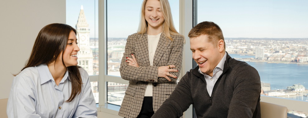 Three individuals smiling and looking at a laptop. The background is a window overlooking a cityscape